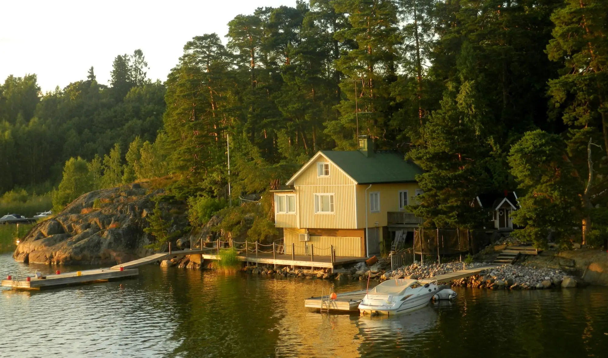 A yellow wooden summer cottage among pine trees on a rocky shore, with a jetty and a moored motorboat on calm water in evening light.