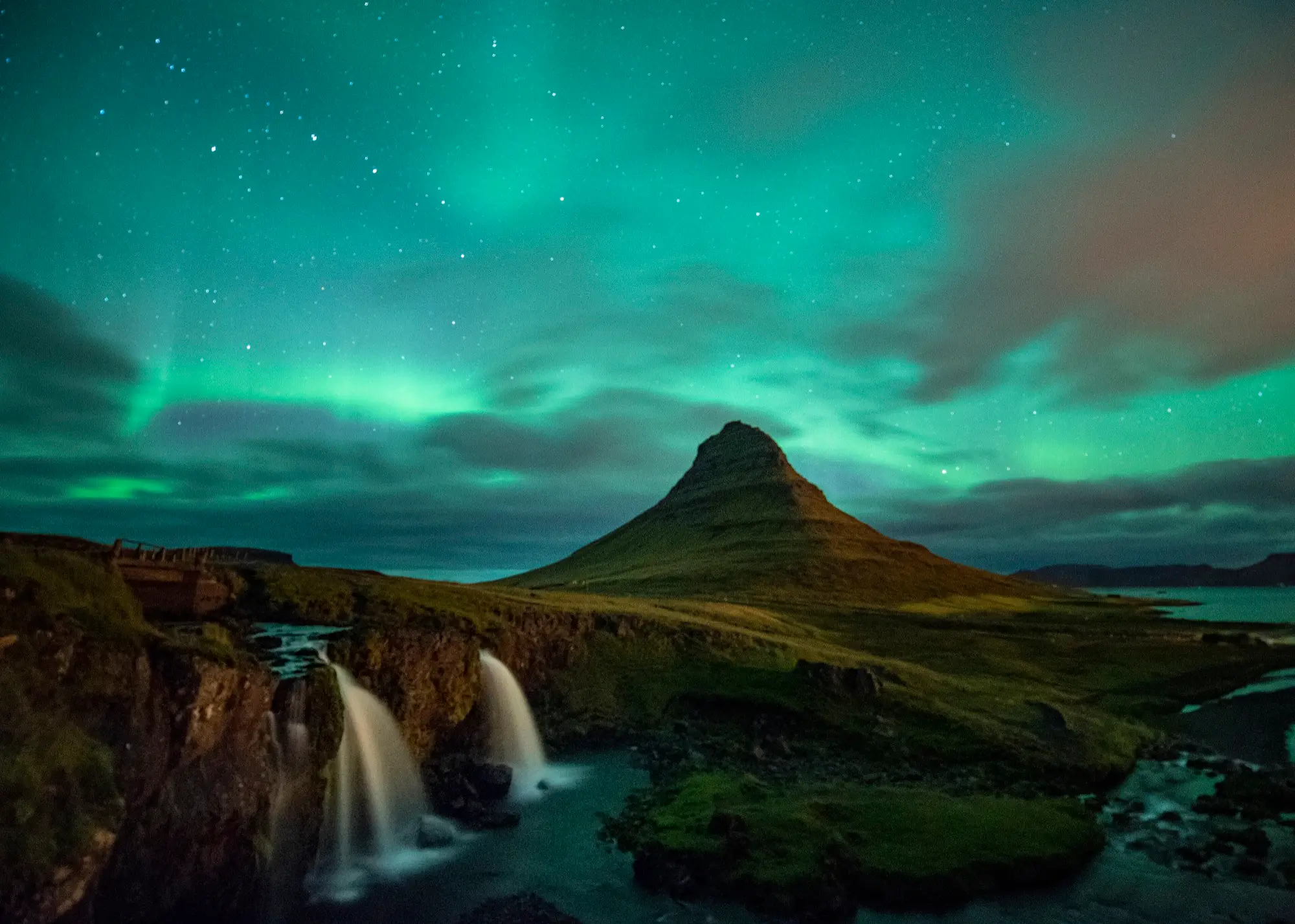 Green aurora over the cone of Kirkjufell mountain on Iceland's Snæfellsnes peninsula, with the Kirkjufellsfoss waterfalls in the foreground.