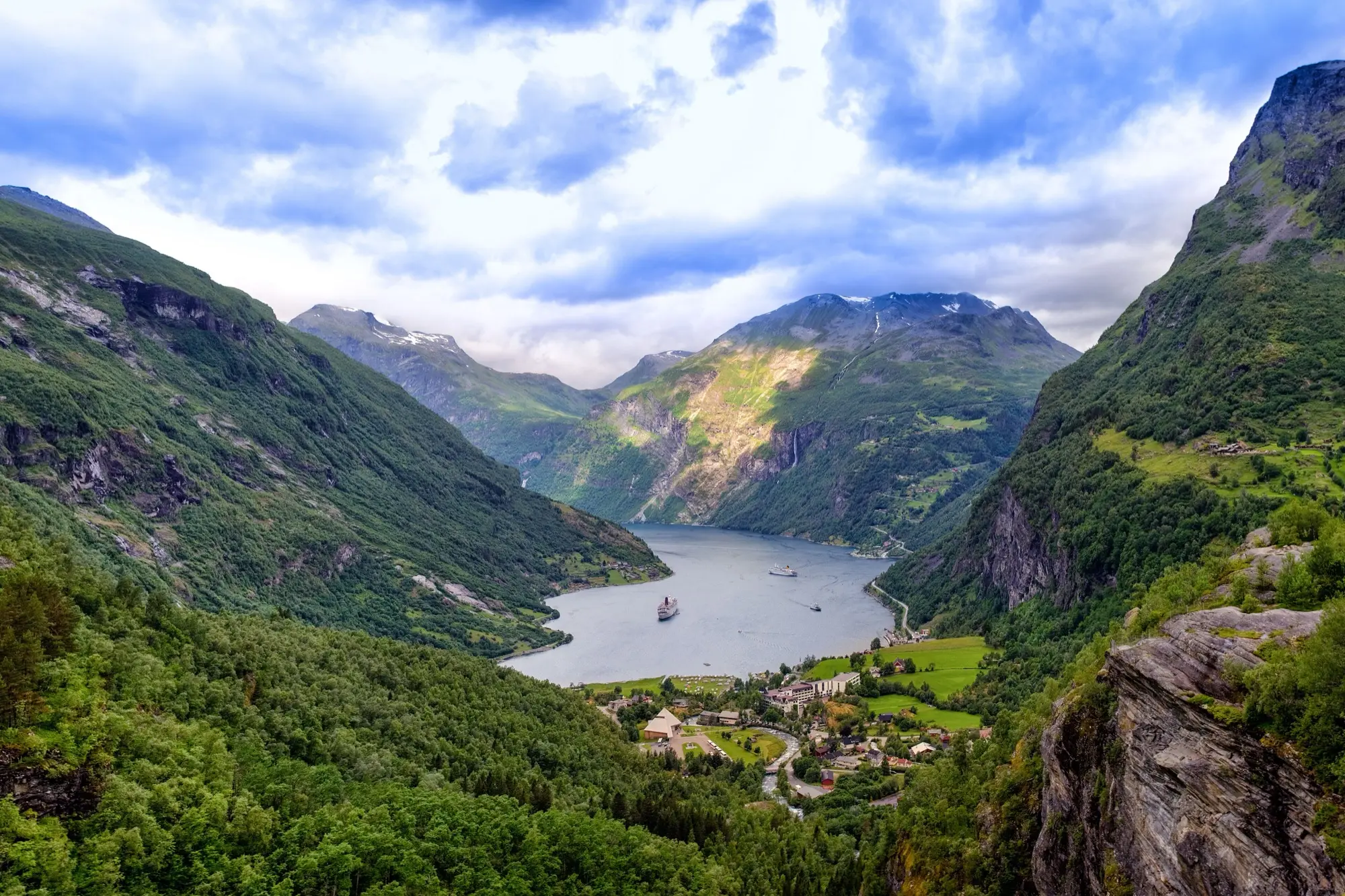 Summer view down the Geirangerfjord from a forested hillside, with green slopes, snow-flecked peaks, boats on the still water and the village of Geiranger in the foreground.