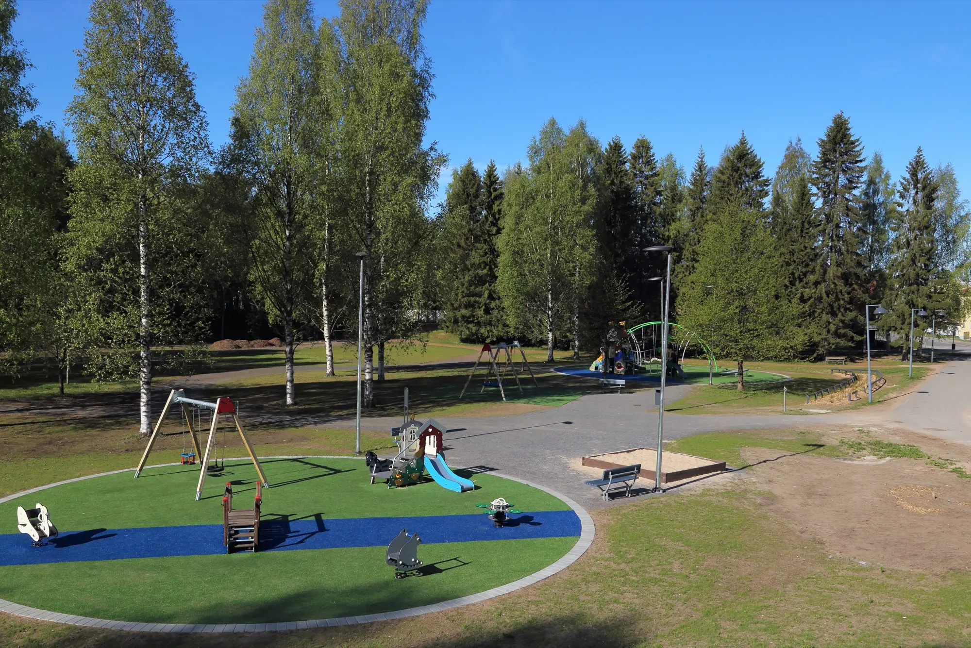 A children's playground with swings, a slide and spring riders on a circle of blue and green surfacing in a sunny birch park in Oulu.
