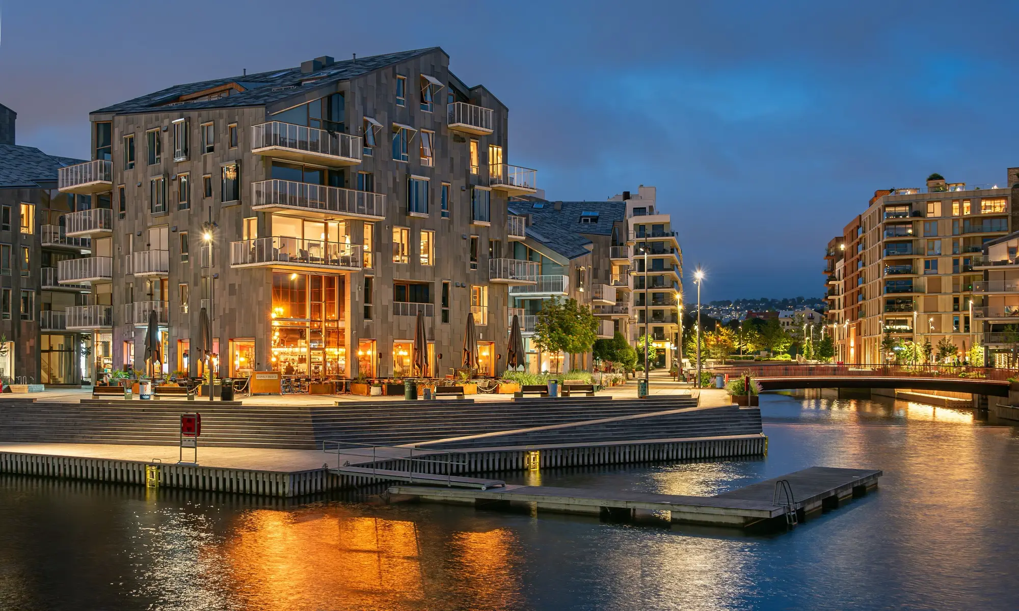 Modern apartment blocks with glass balconies line a canal-side waterfront in Bjørvika, Oslo, their windows and ground-floor cafés lit up at blue hour and reflected in the water.