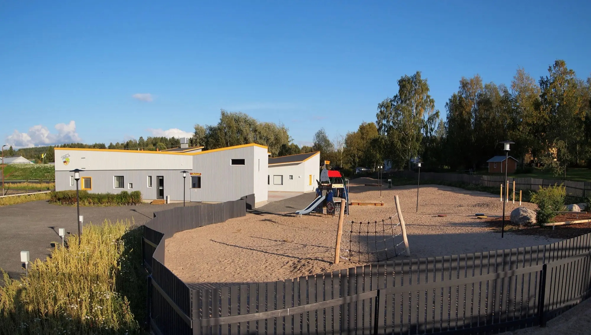 A low white daycare building with a fenced sandy playground yard and climbing frames under a blue sky in Jyväskylä.