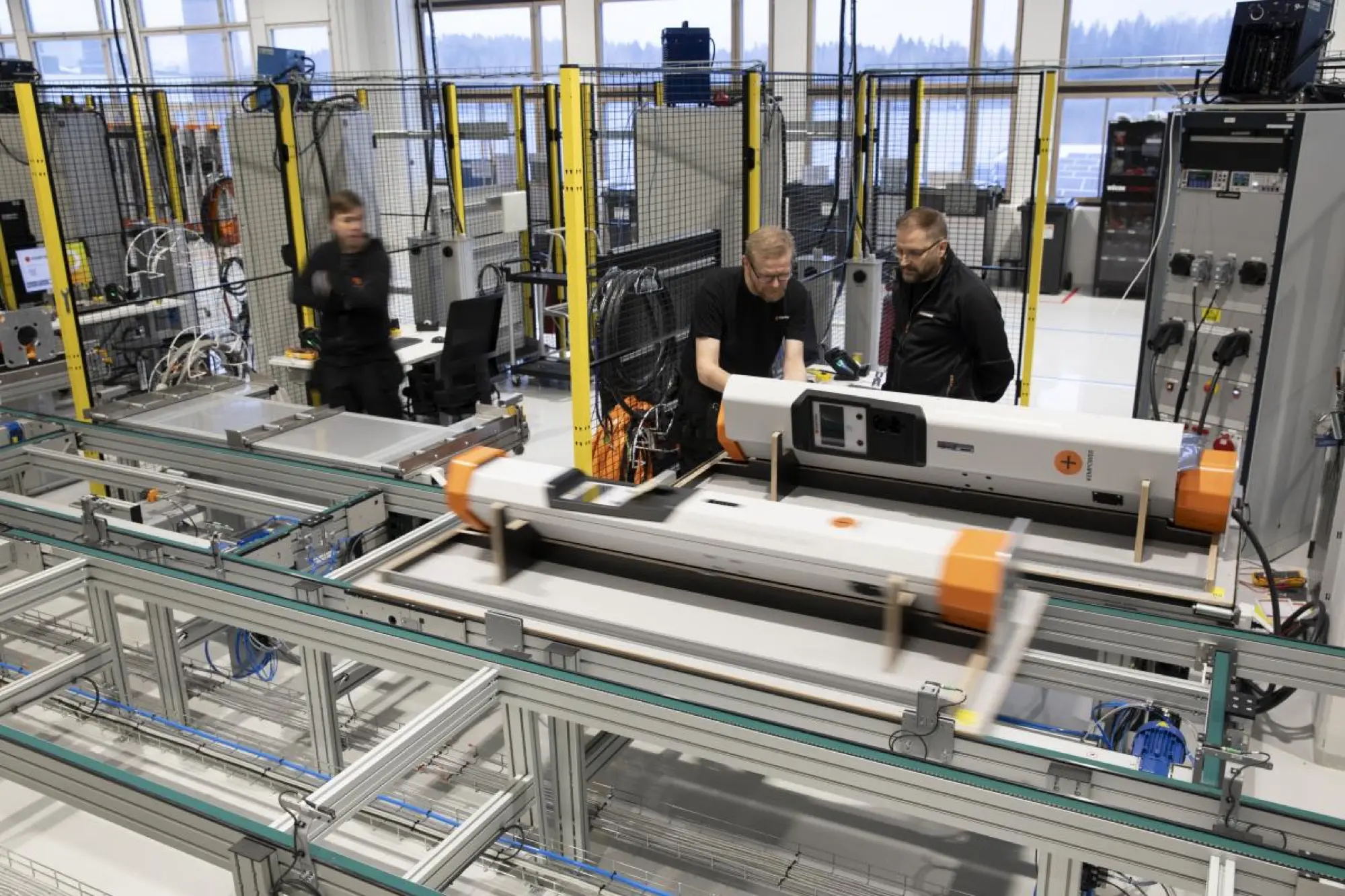 Workers testing electric vehicle chargers at workstations on a bright factory floor in Lahti.