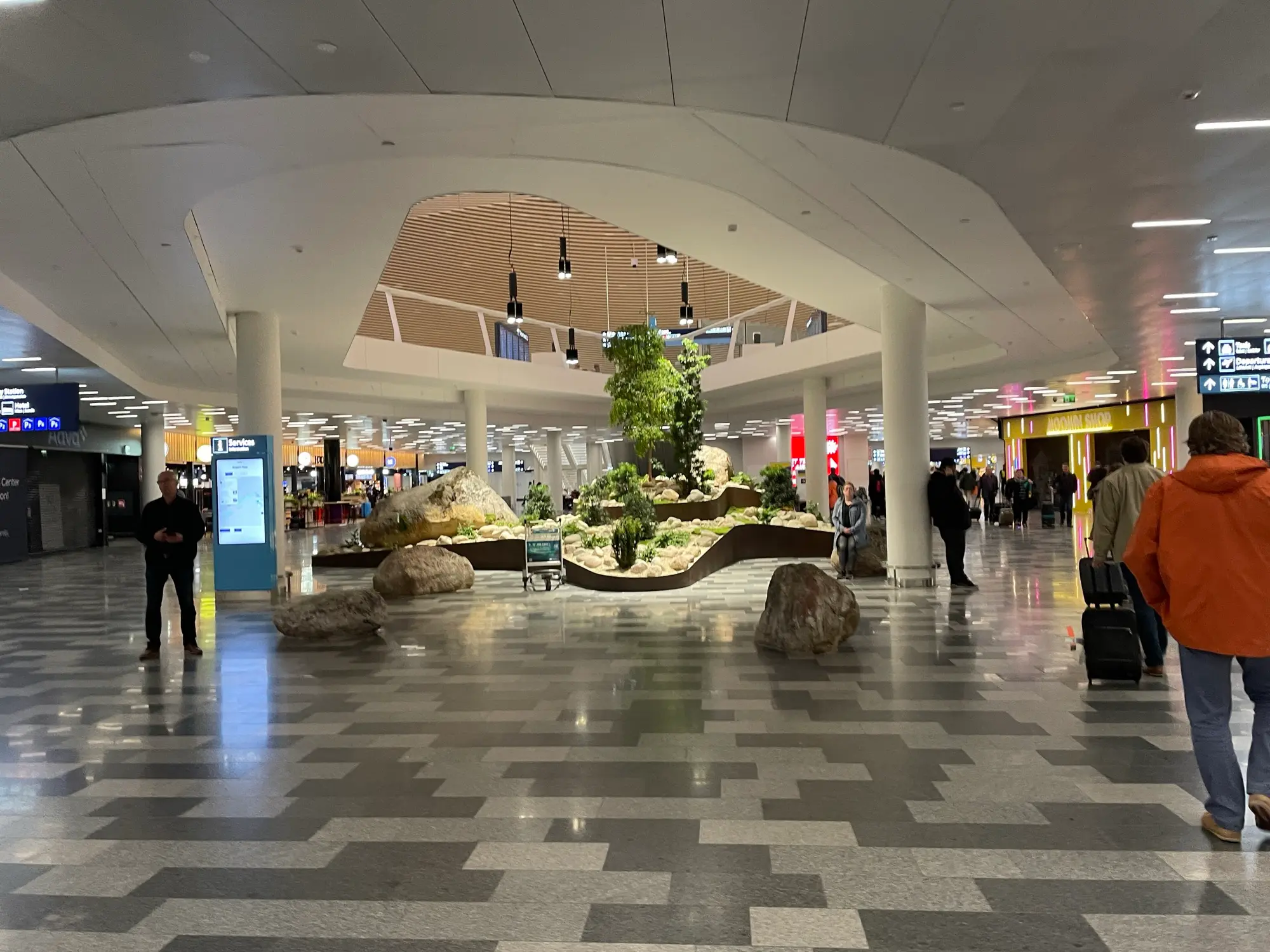 The renovated arrivals floor of Helsinki Airport's Terminal 2, with travellers walking past a planted rock garden and round white columns.