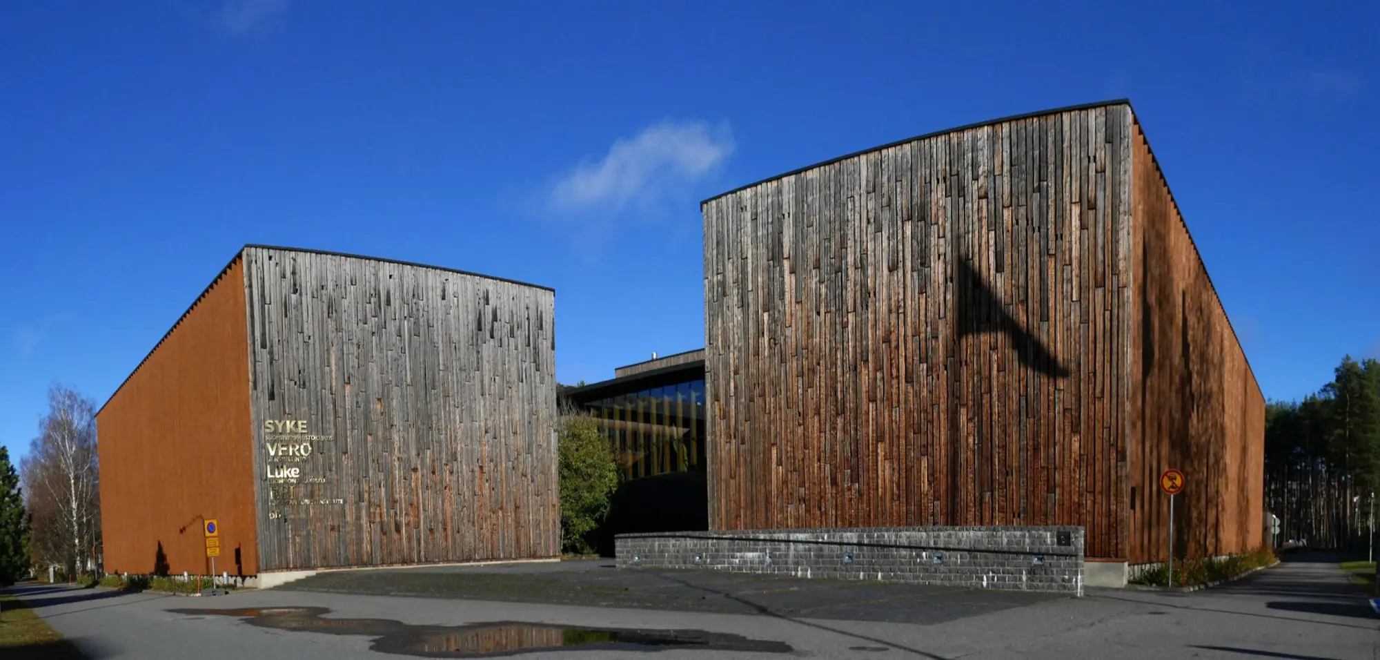 Two tall timber-clad government office buildings in Joensuu, one with SYKE, VERO and LUKE lettering on its wooden facade, under a deep blue sky.