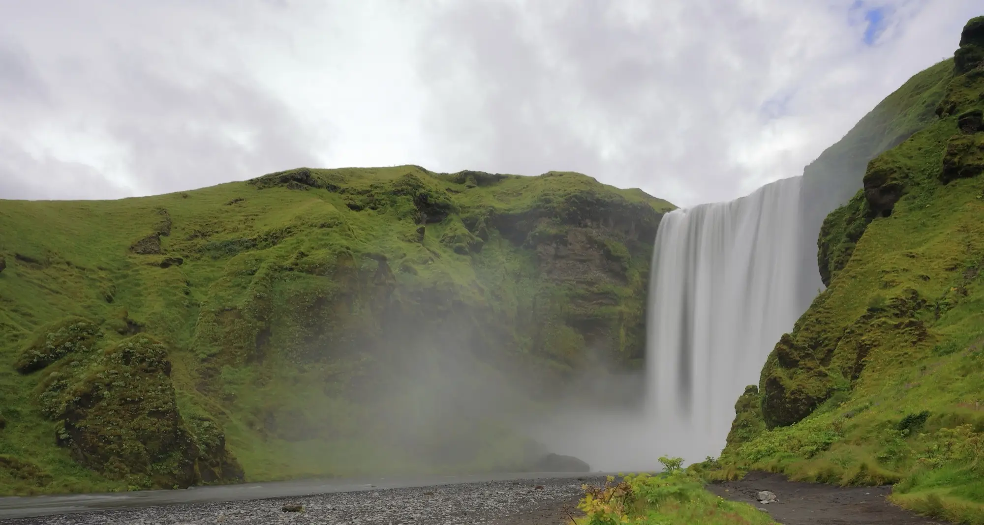 The Skógafoss waterfall dropping from a green cliff in south Iceland, with spray drifting across the meadow below.