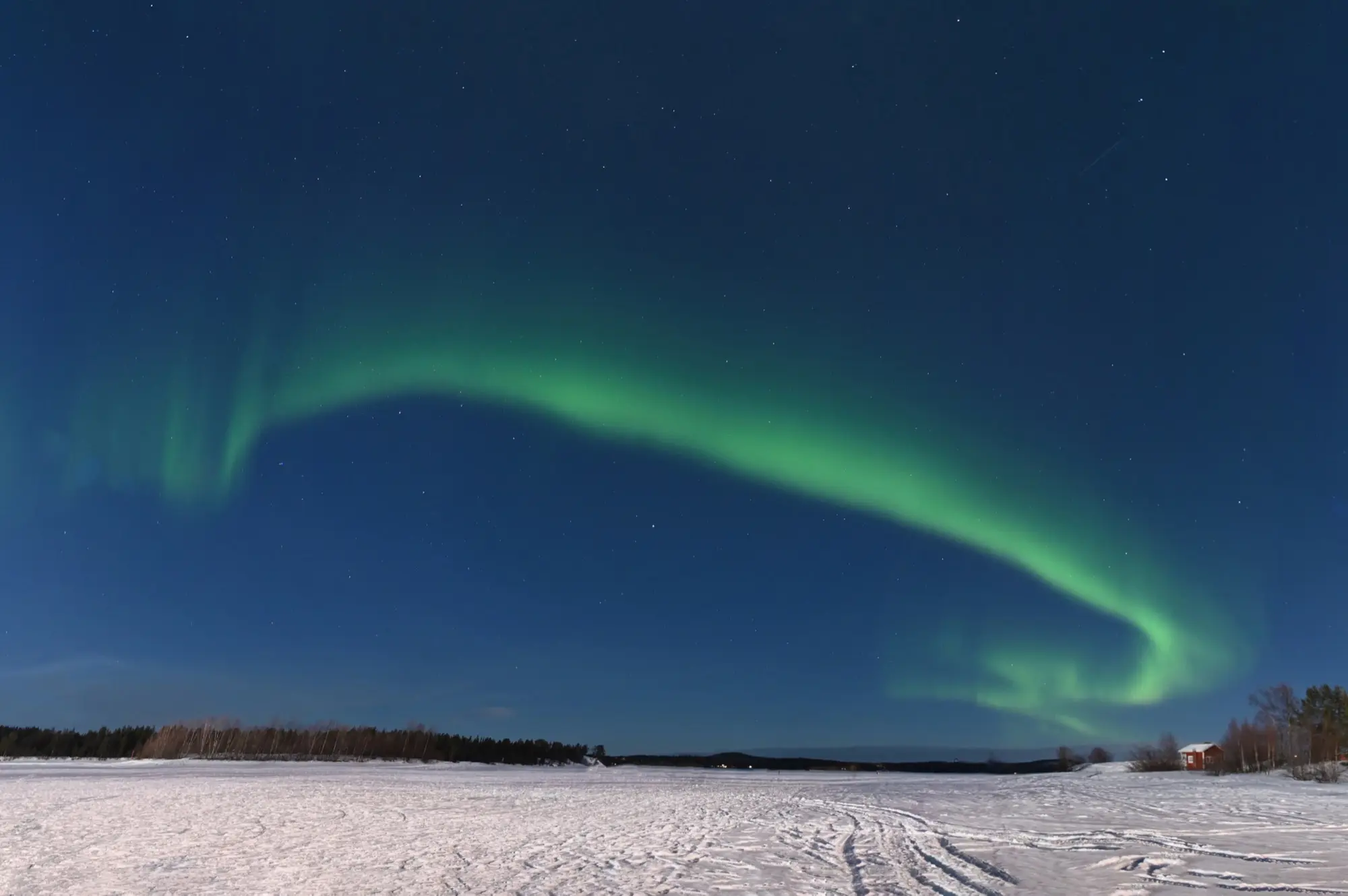 A long green arc of northern lights across a starry blue sky at Inari in March, above a frozen, snow-covered lake marked with tracks, a line of forest and a small red cabin on the shore.