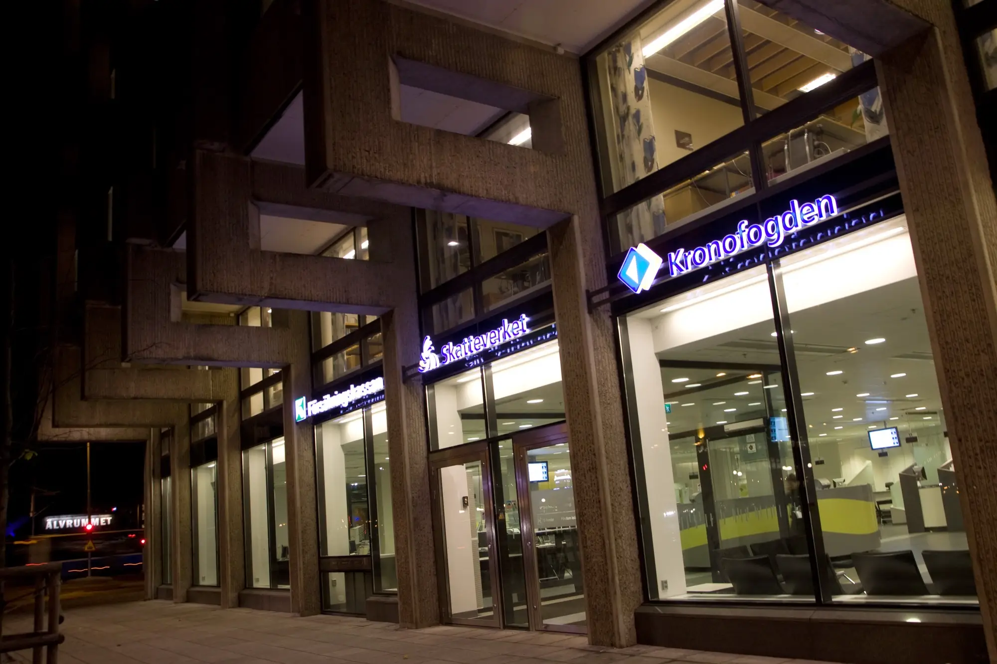 The glass entrance of a government service office in Gothenburg at night, with lit Skatteverket and Kronofogden signs on a concrete facade.