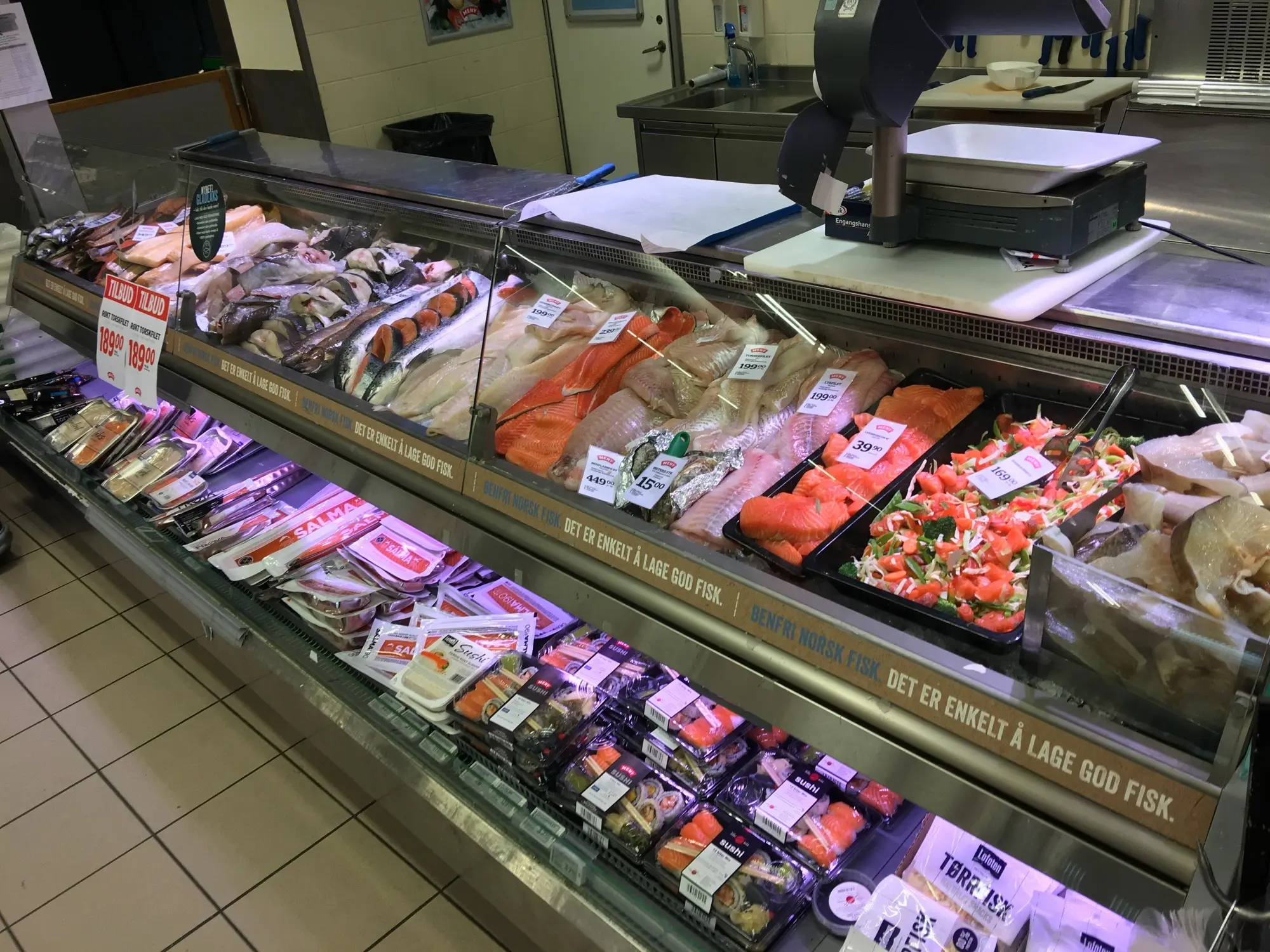 A supermarket fish counter in Bergen with salmon steaks, fillets, ready-made sushi and seafood salads on ice, each labelled with kroner price tags and a red discount sign.