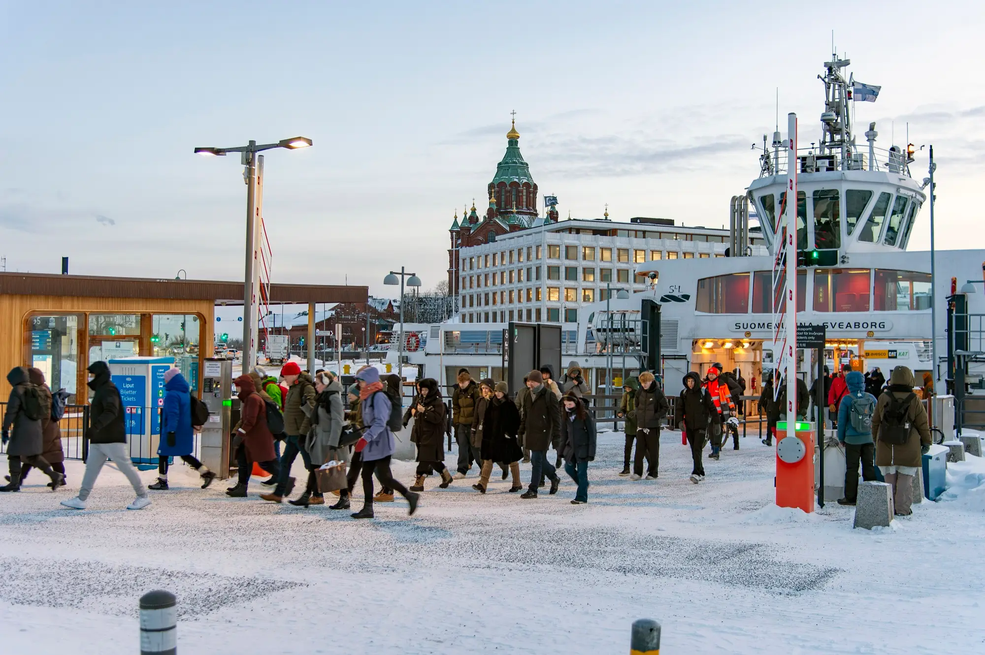 Commuters in winter coats walk off a ferry across the snowy Market Square quay in Helsinki on a grey morning.