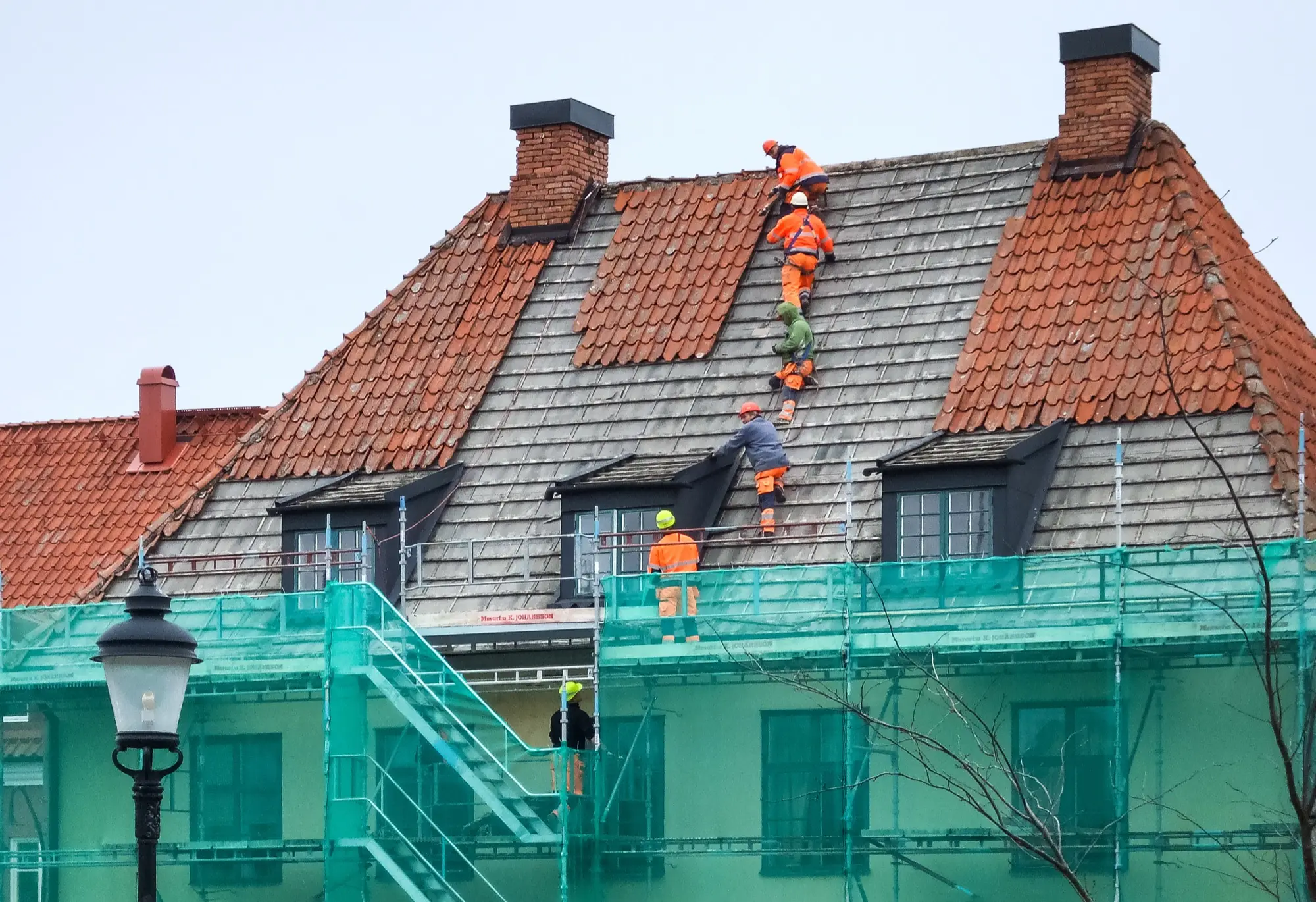 Roofers in orange high-visibility workwear replacing tiles on a steep house roof above scaffolding in Visby, Gotland.
