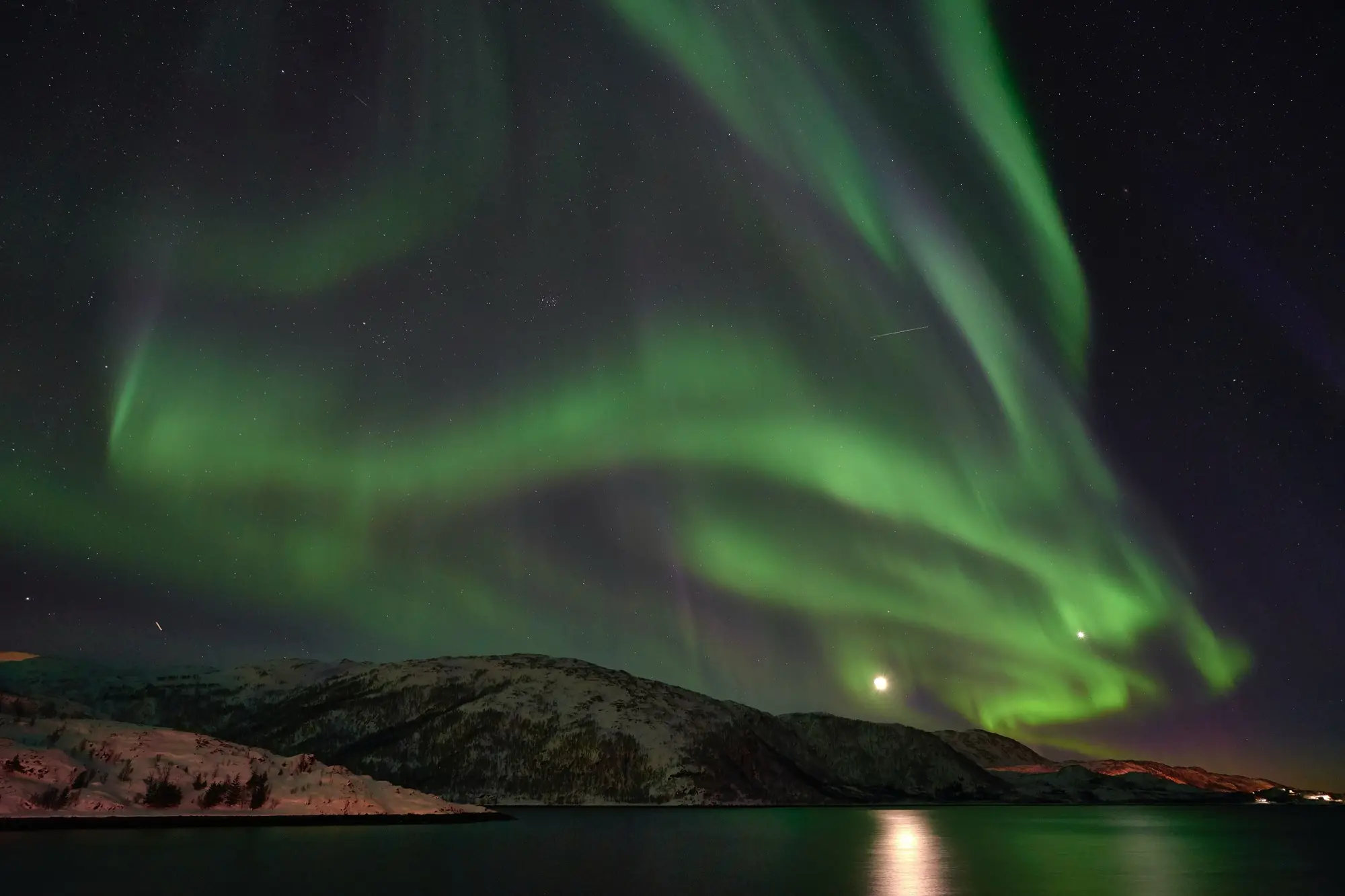 Bright green northern lights swirl across a starry sky above snow-dusted hills and a calm fjord at Lauklines on Kvaløya, near Tromsø, with the moon reflected in the water.