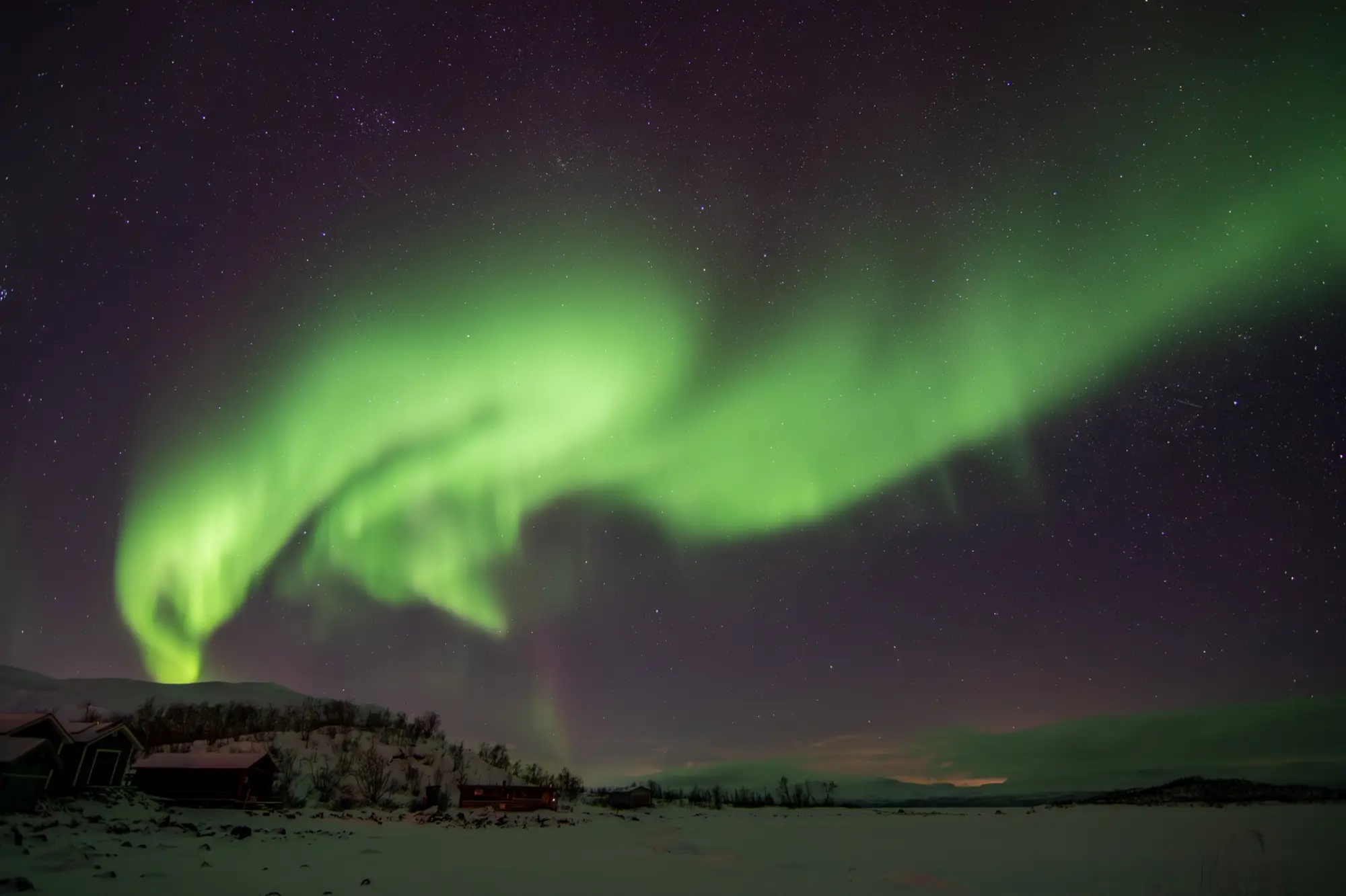 A swirl of green northern lights over red wooden cabins and a snow-covered plain in Abisko National Park, Swedish Lapland.