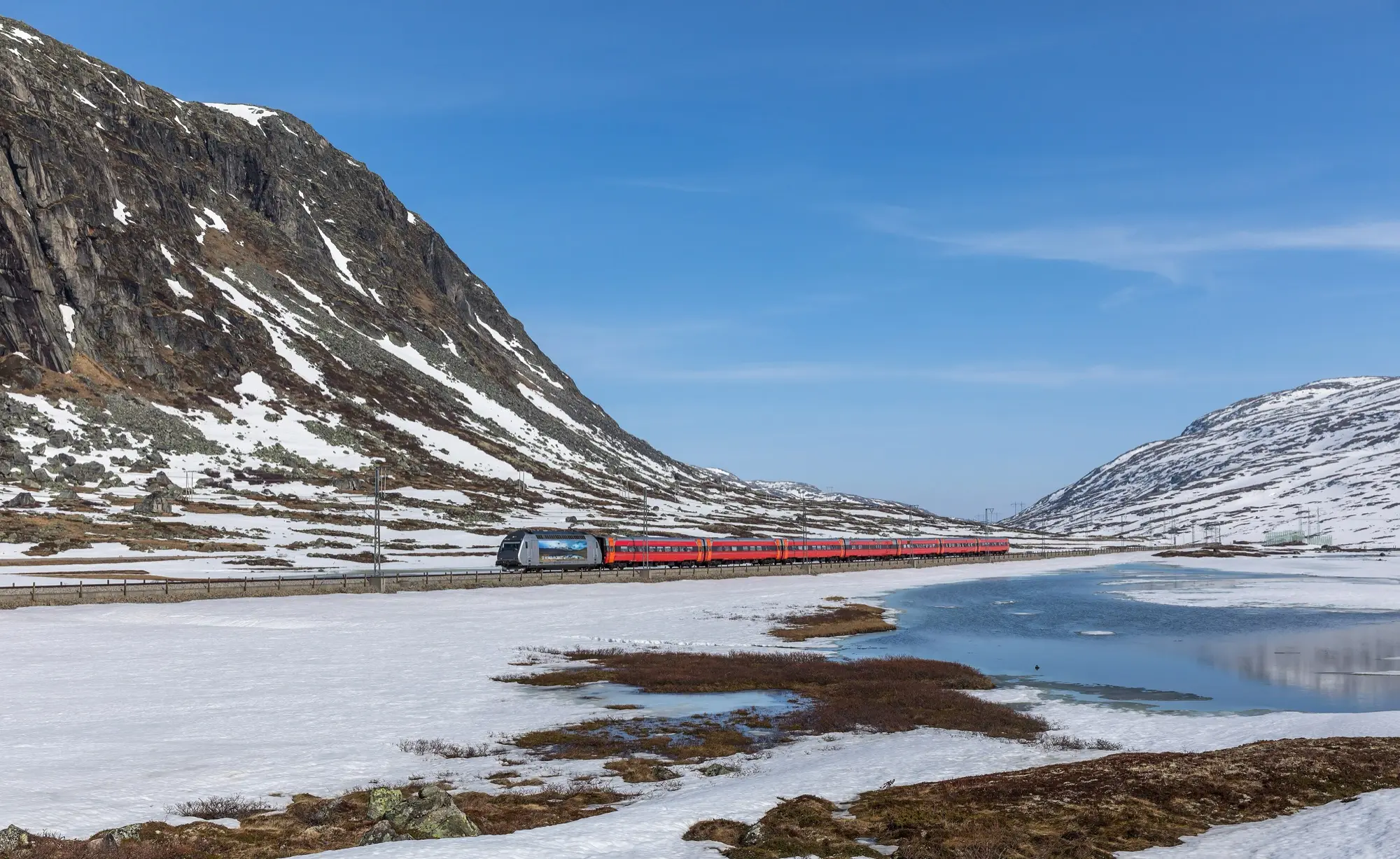 A red Vy Bergen Line train hauled by a silver El 18 locomotive crosses the snowy Hardangervidda plateau between Haugastøl and Finse, beside a half-frozen lake under a clear blue sky.