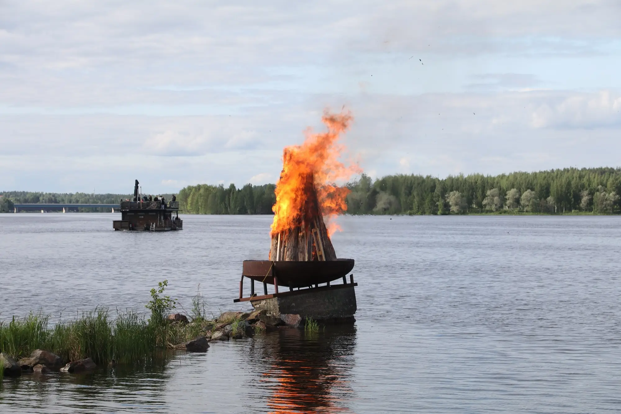 A Midsummer bonfire burning on a floating platform on the water in Imatra, with a forested shore behind.