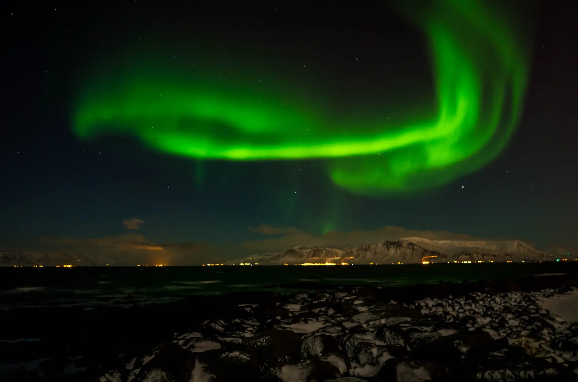 A bright green aurora curving over the bay at Reykjavík in winter, with the snow-covered Esja mountain and a line of shore lights across the water and snowy rocks in the foreground.