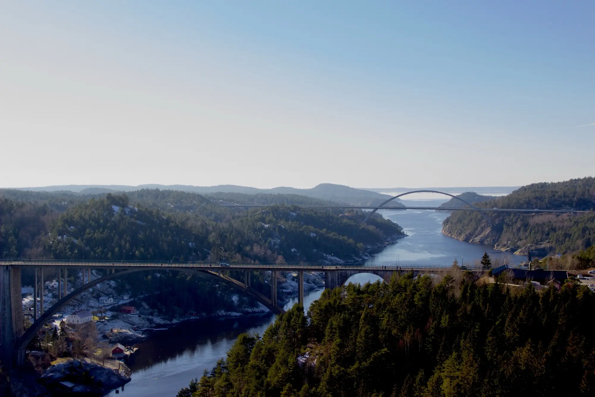 The old Svinesund arch bridge in the foreground and the new white-arched Svinesund bridge beyond it, both crossing the forested Iddefjord on the border between Sweden and Norway.