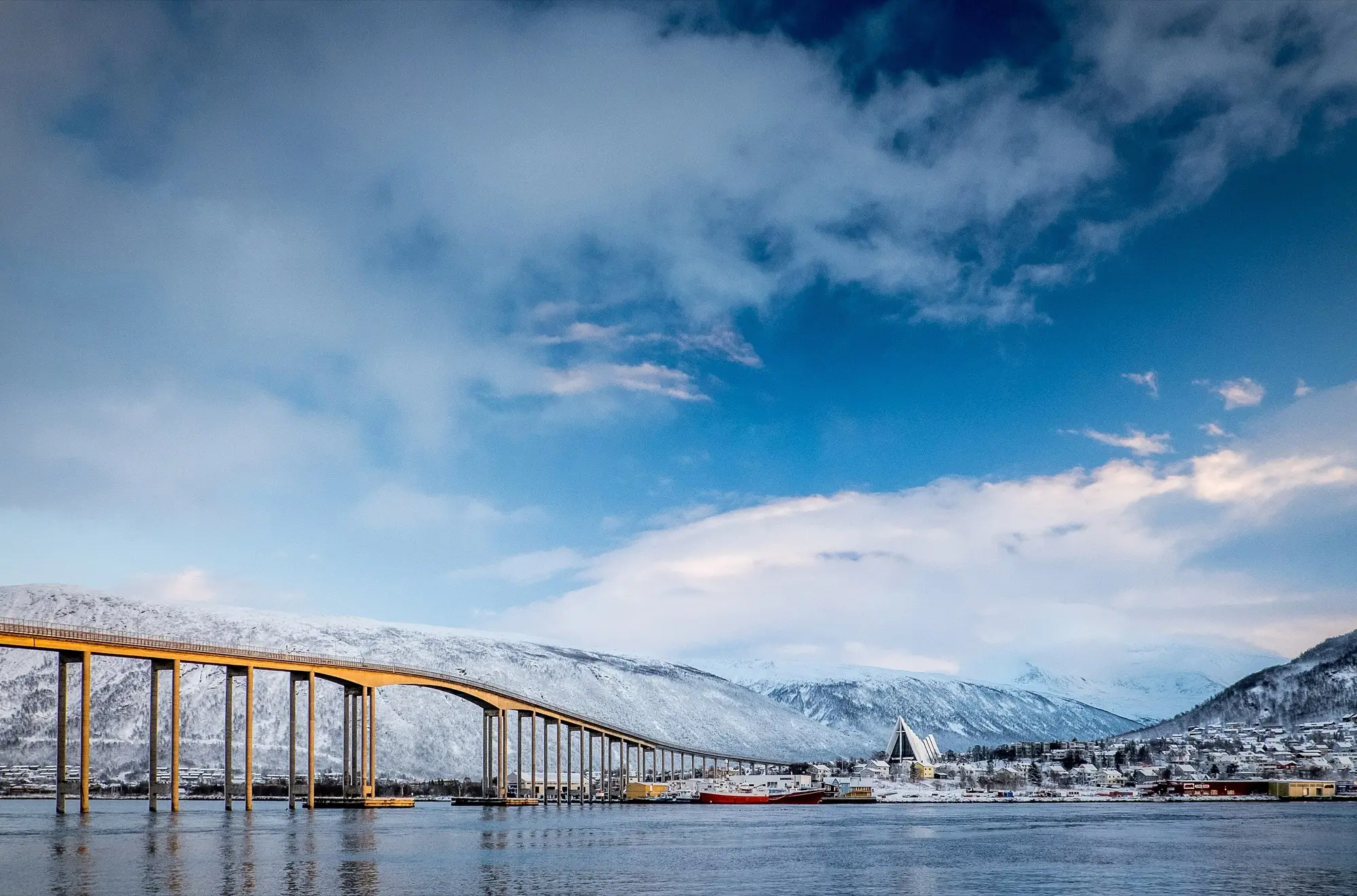 The long arched Tromsø Bridge spans the sound towards the white, triangular Arctic Cathedral and snow-covered Tromsdalen, with snowy mountains and a cold blue winter sky behind.