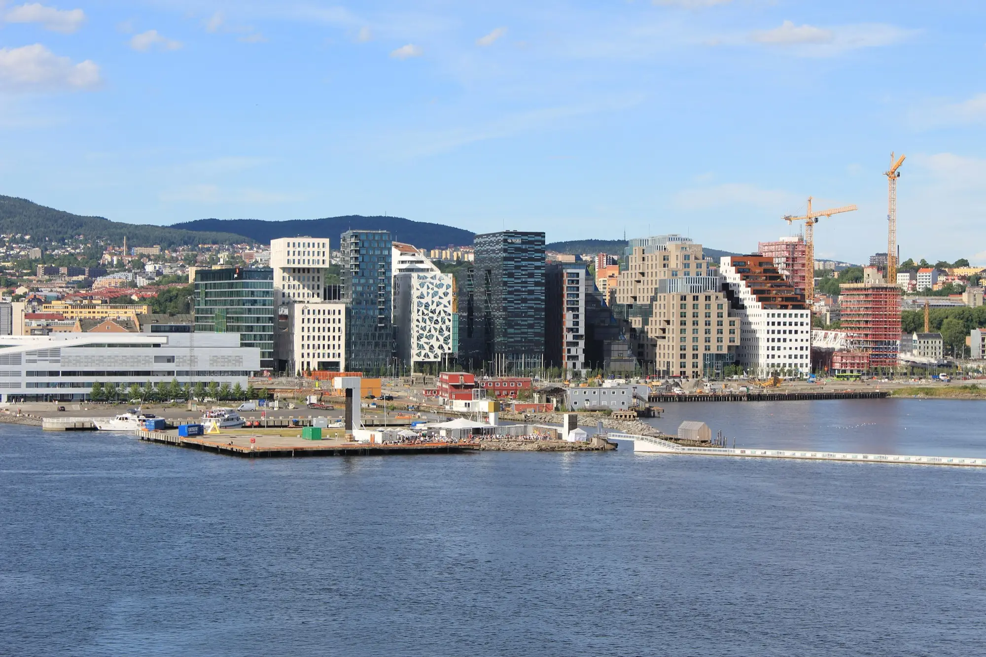 The Barcode row of glass and patterned office towers in Bjørvika, Oslo, seen across the harbour on a sunny day, with construction cranes and the forested hills behind the city.